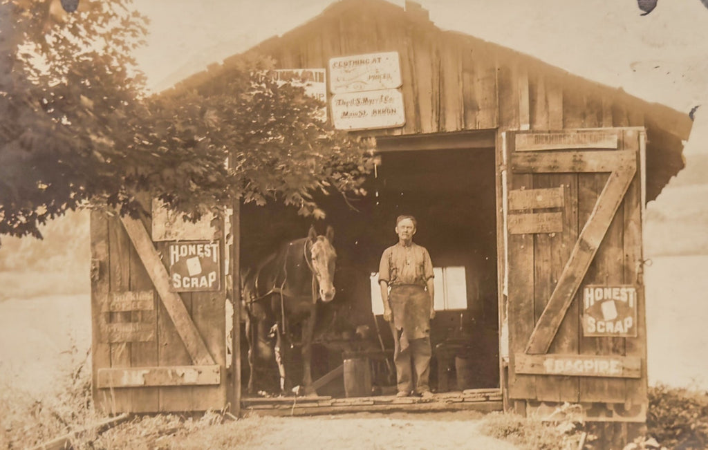 Vintage sepia-toned photograph of a man standing in front of a wooden shed with a horse inside.