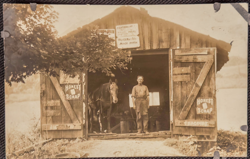 Vintage sepia-toned photograph of a man standing in front of a wooden shed with a horse inside.