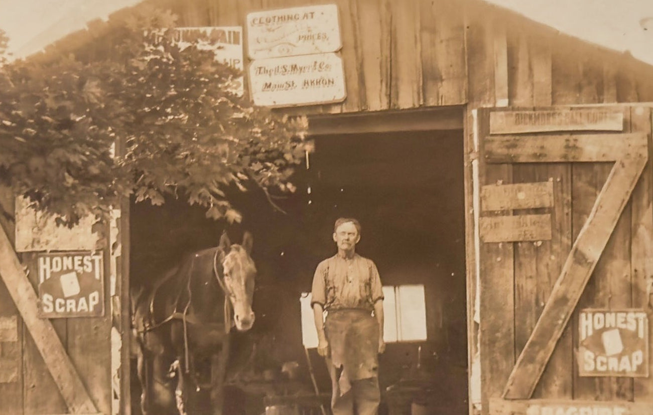 Vintage sepia-toned photograph of a man standing in front of a wooden shed with a horse inside.