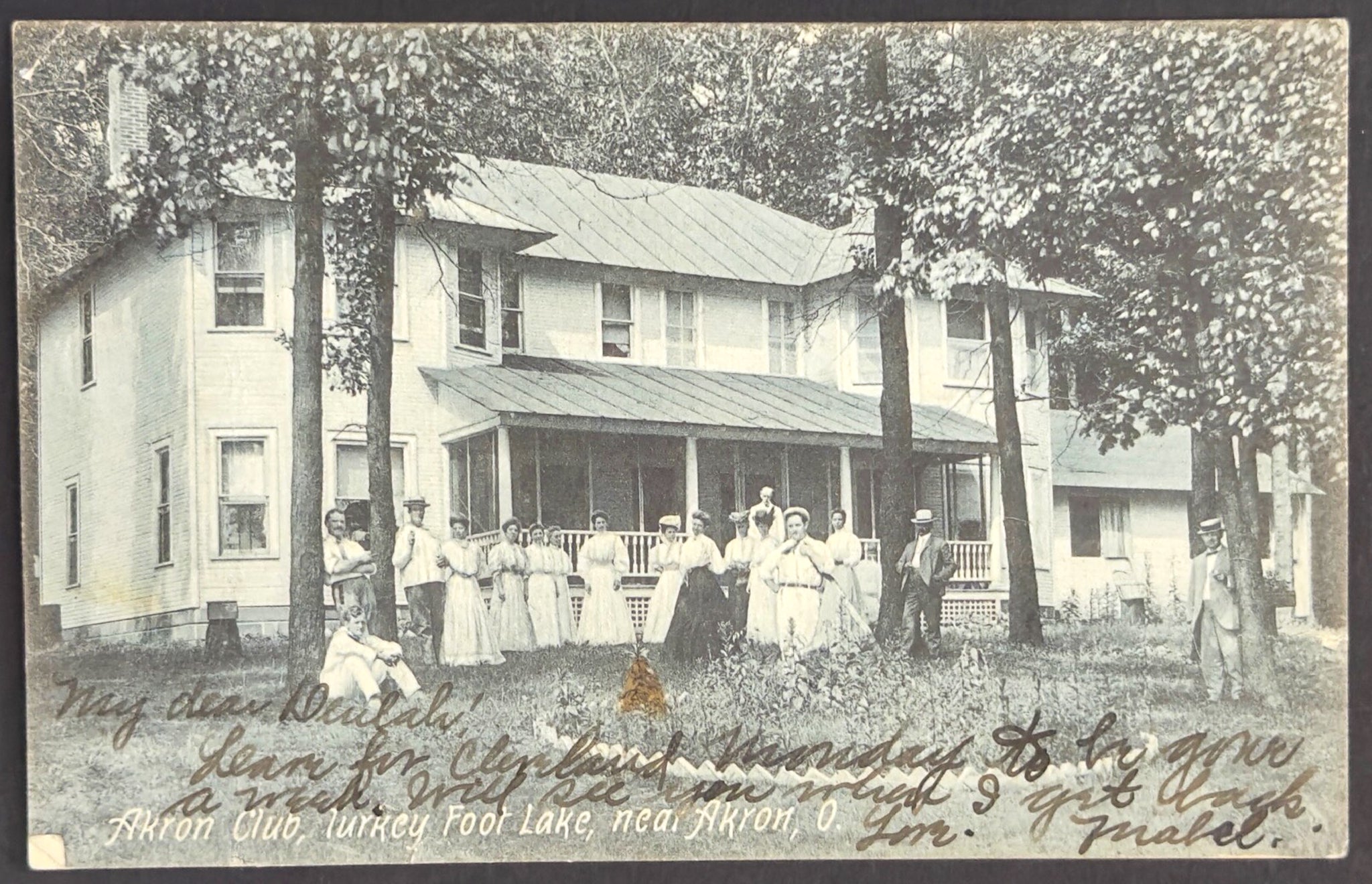 Vintage photograph of a group of people standing on a porch in front of a house with trees and a garden.