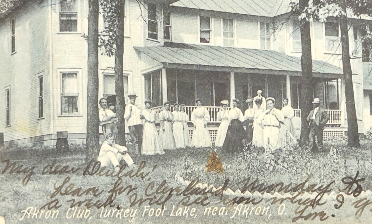 Vintage photograph of a group of people standing on a porch in front of a house with trees and a garden.