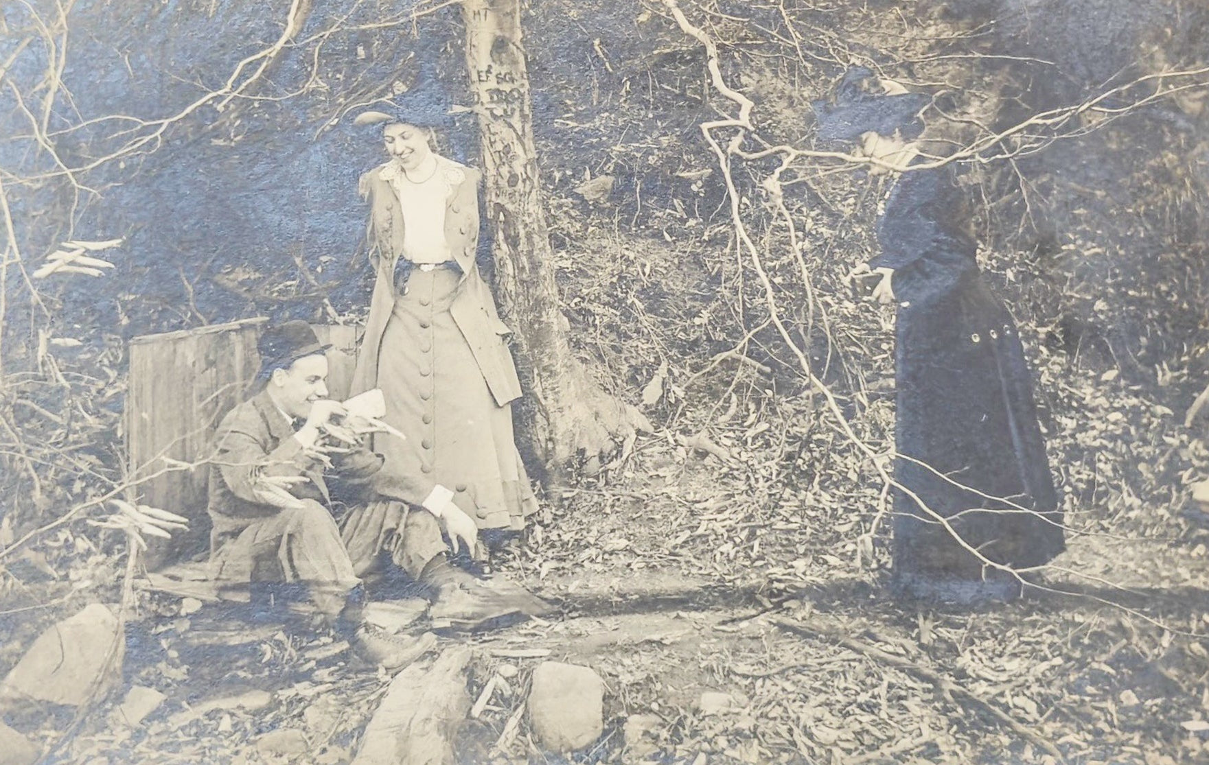 Vintage photograph of people in a forest setting with a woman in a dress and two others.