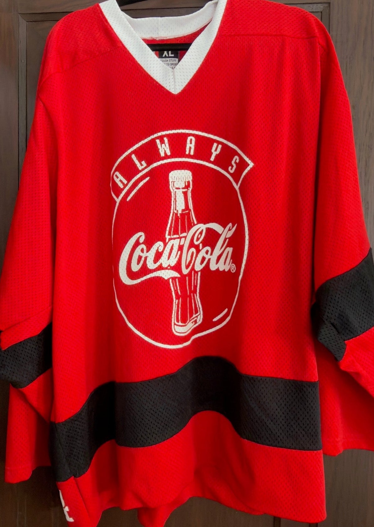 Red jersey with Coca-Cola logo on a hanger against a wooden door.