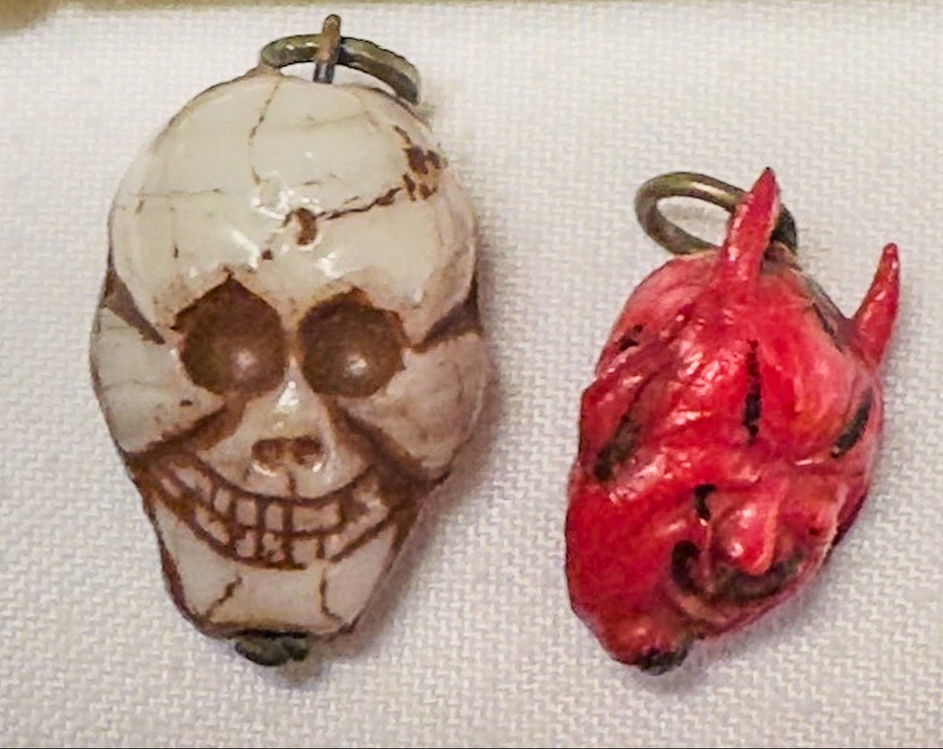 Two small pendants, one skull-like and one red, next to a measuring tape on a white background.